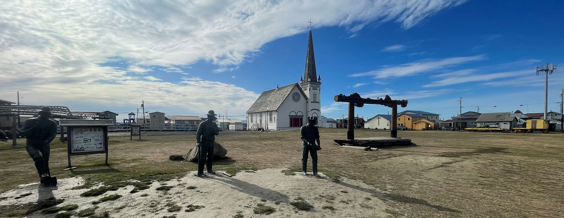 Statues and the Iditarod finish line are pictured in the foreground. A church and other buildings in the city of Nome, Alaska are pictured in the background.