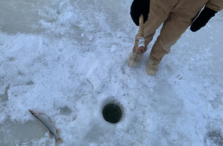 A person stands on the ice holding an ice fishing rod, extending to a hole in the ice. A small fish lays on the ice.