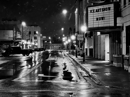 A black and white photo of downtown Fairbanks where the bright city lights before sunrise highlight a light snowfall and wet city streets
