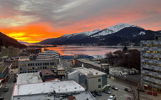 The sun rises behind snow capped mountains, turning the sky orange and red. Buildings of the city of Juneau in the forgraound.