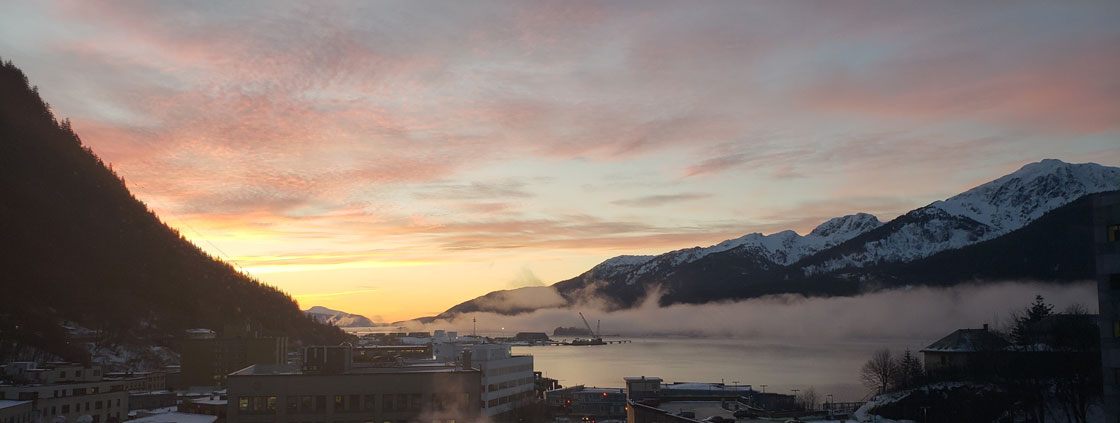 Sunrise colors the clouds orange and pink behind the mountains with the city of Juneau below as seen from out the Juneau office window 11/8/19.