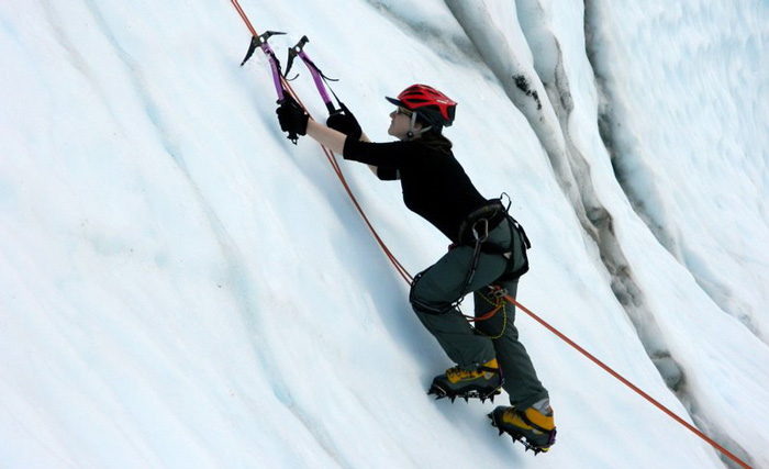Bailey Woolstead wearing ice climbing gear, climbs up a wall of ice.